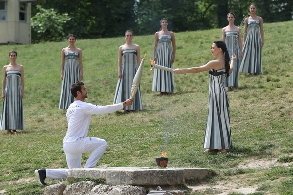 扮演最高女祭司的演員瑪麗·米娜（前右）點(diǎn)燃第一棒火炬手——希臘運(yùn)動員、東京奧運(yùn)會男子賽艇項(xiàng)目金牌得主斯特凡諾斯·恩圖斯科斯手中的火炬。新華社記者 趙丁喆 攝